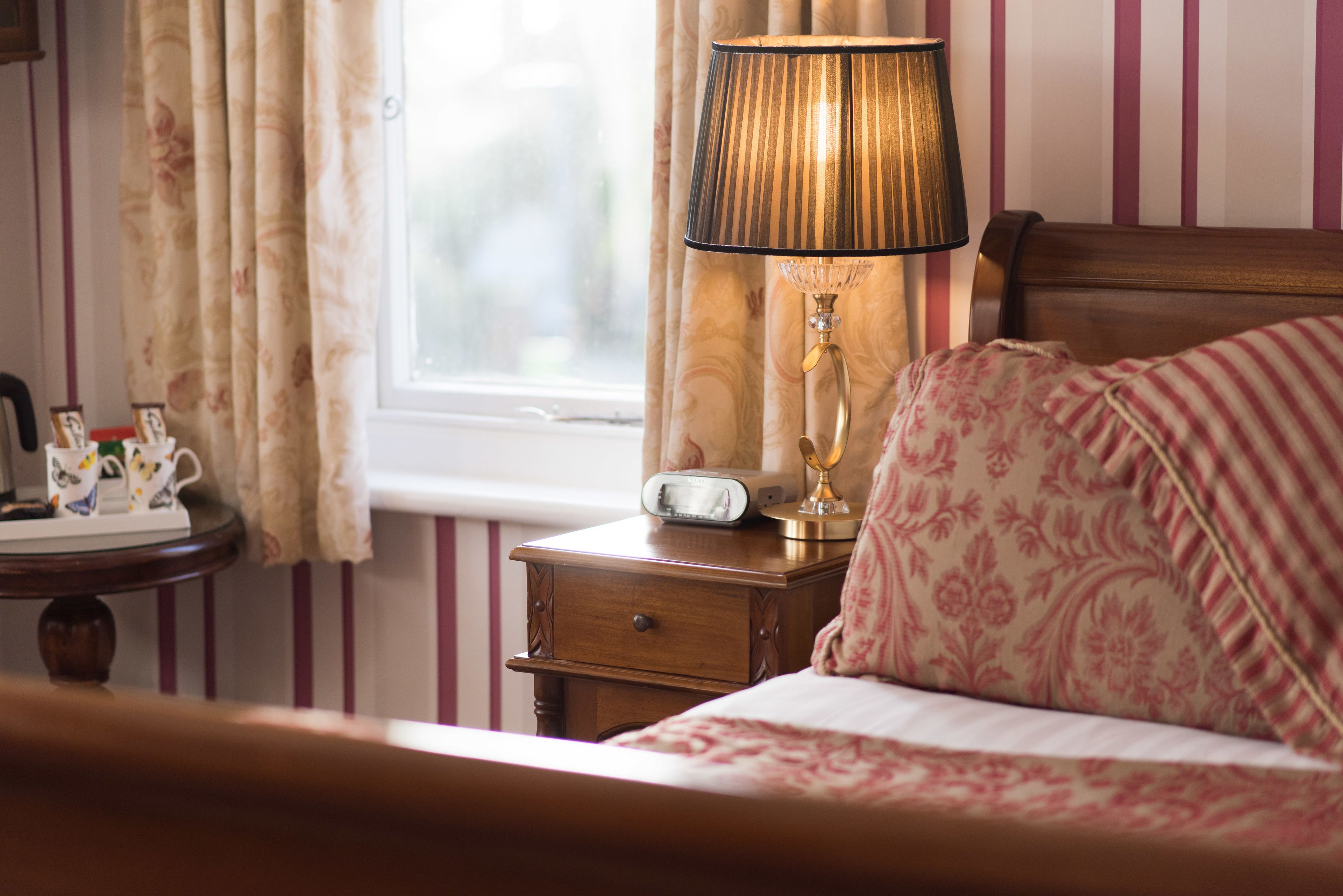 A bedroom with a bed, a bedside table with a lamp, a clock, and a window with curtains.
