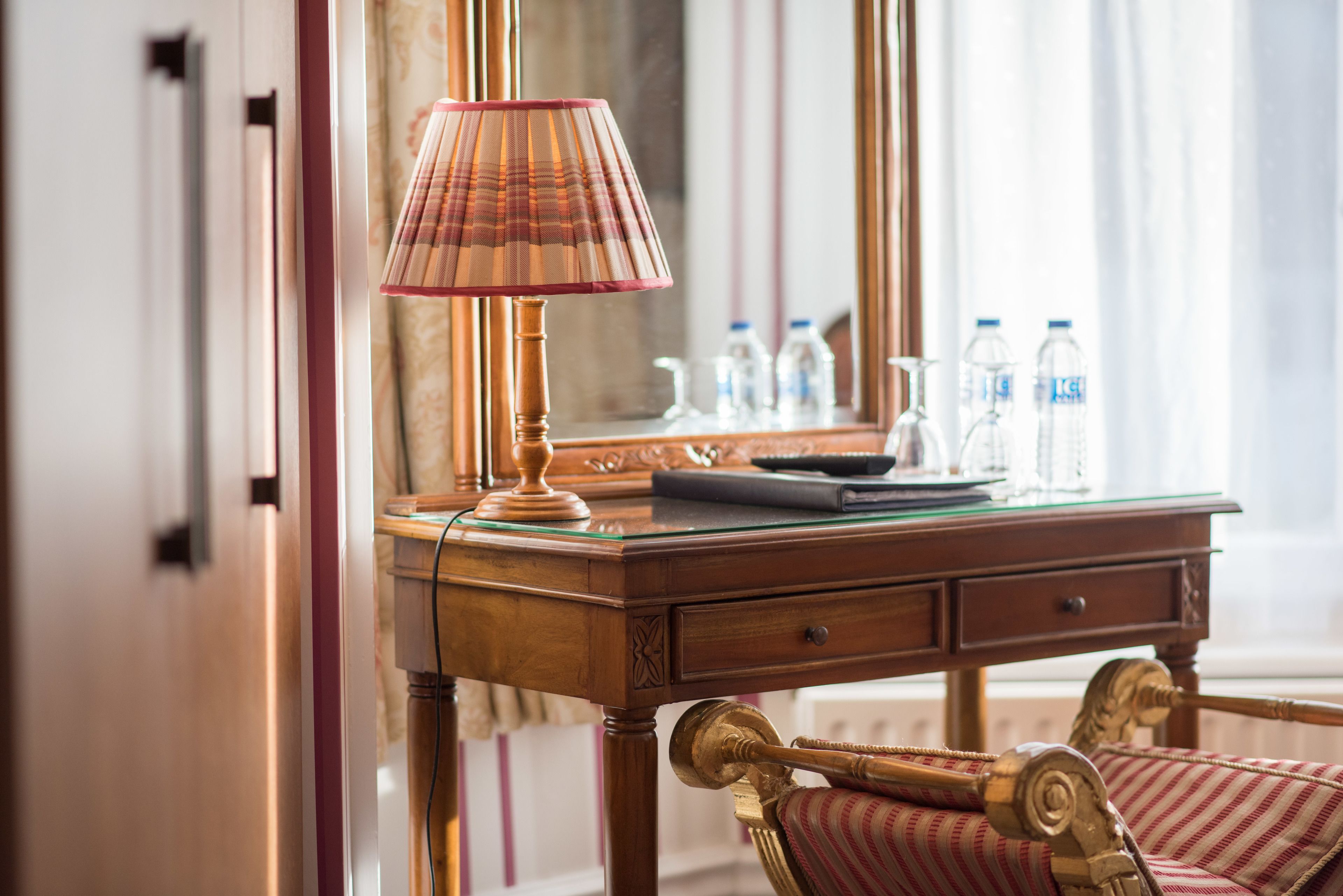 A wooden desk with a lamp, a mirror, and a chair. There are water bottles on the mirror.