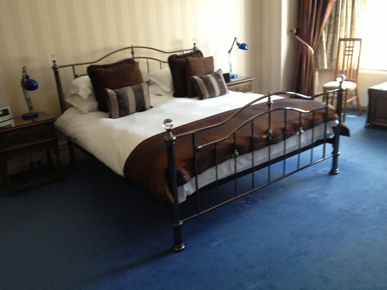 A neatly made bed with a metal frame, a blue carpet, and a bedside table with a lamp.
