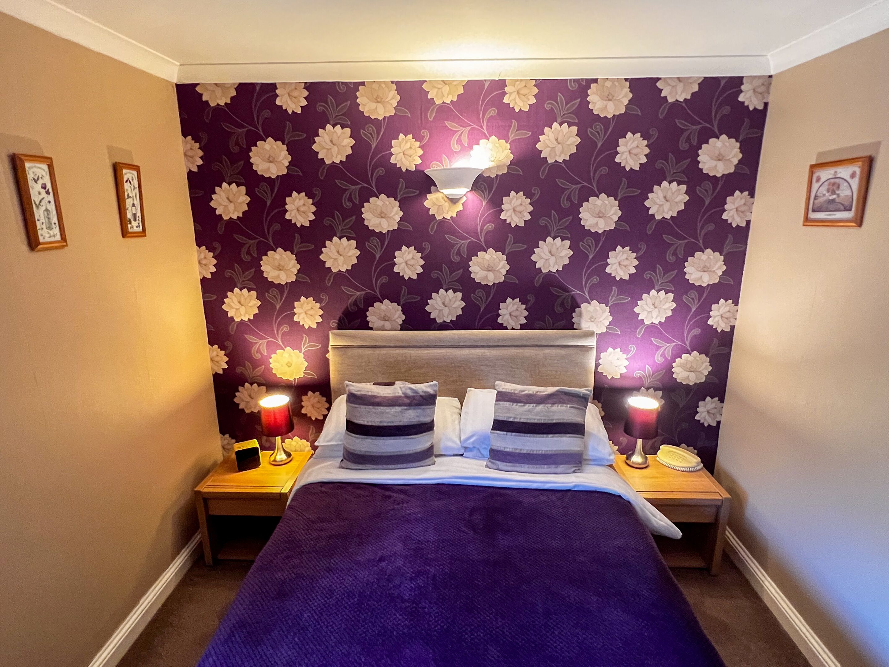 A bedroom with a purple floral wallpaper, a bed with two pillows, two bedside tables with lamps, and framed pictures on the walls.