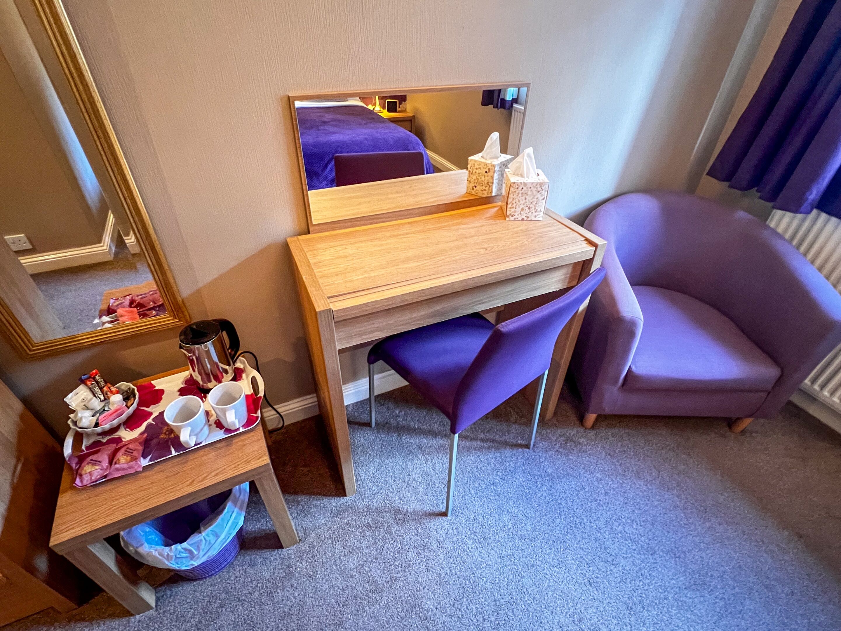 A hotel room with a wooden desk, a purple chair, a small table with a tray of snacks, a mirror, and a radiator.