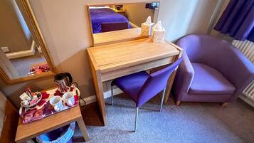 A hotel room with a wooden desk, a purple chair, a small table with a tray of snacks, a mirror, and a radiator.