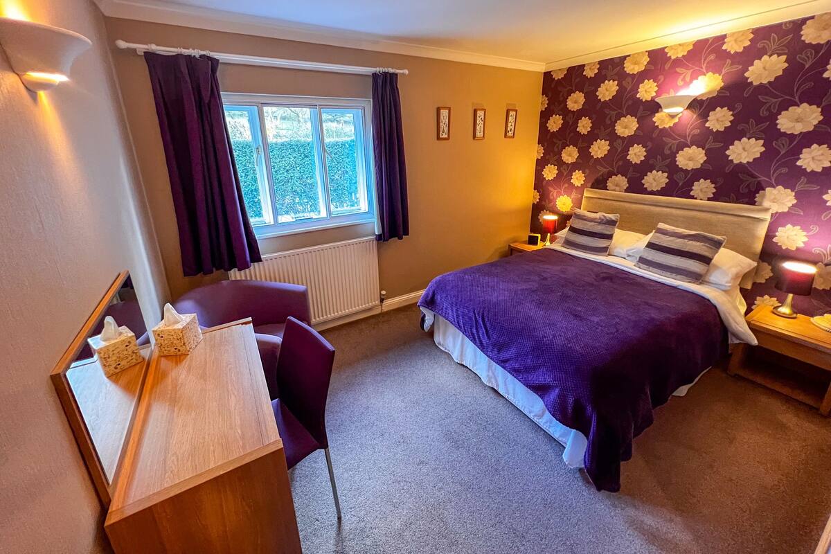 A bedroom with a bed, a desk, a chair, a window with curtains, and a wall with floral wallpaper.