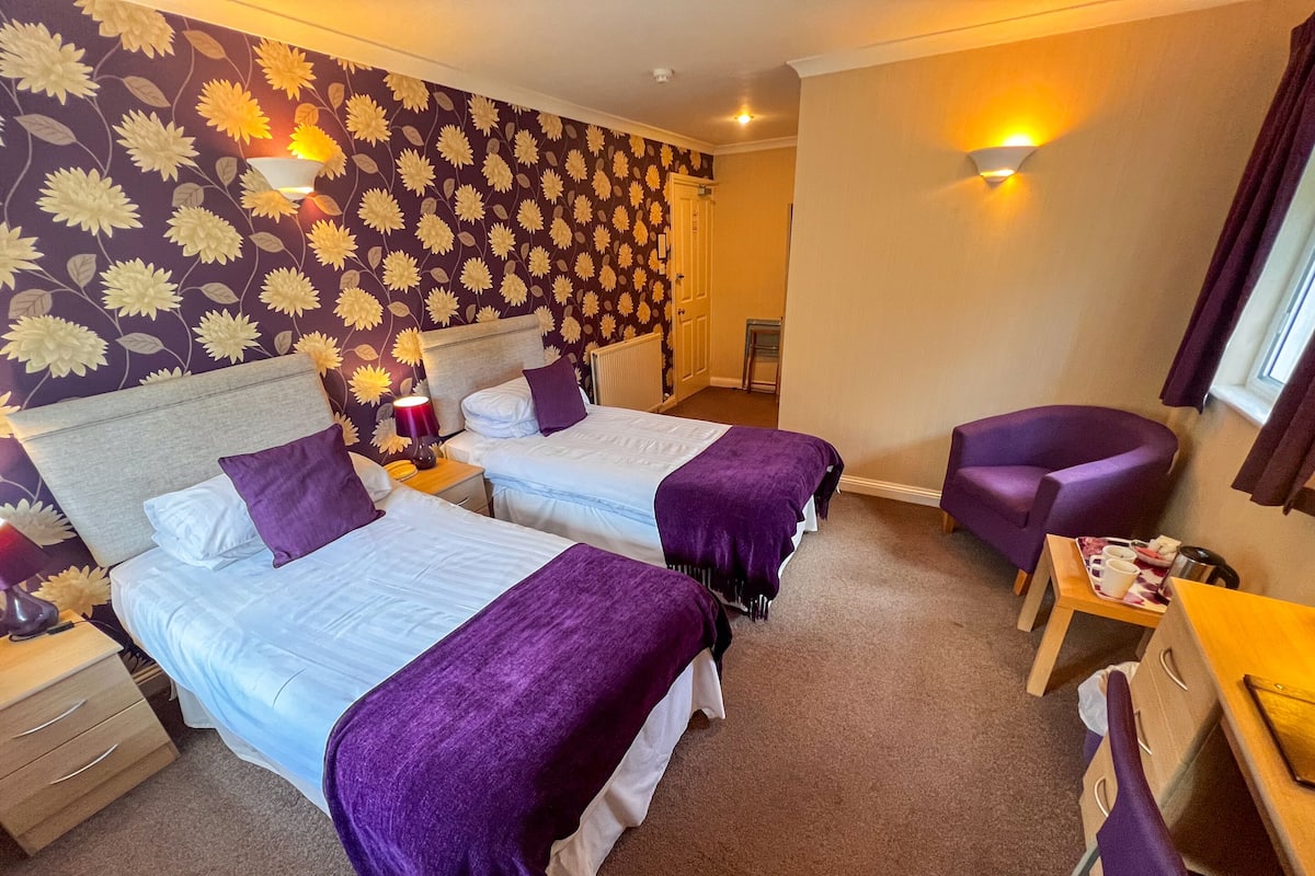 A hotel room with two beds, a desk, a chair, and a wall with a floral pattern.
