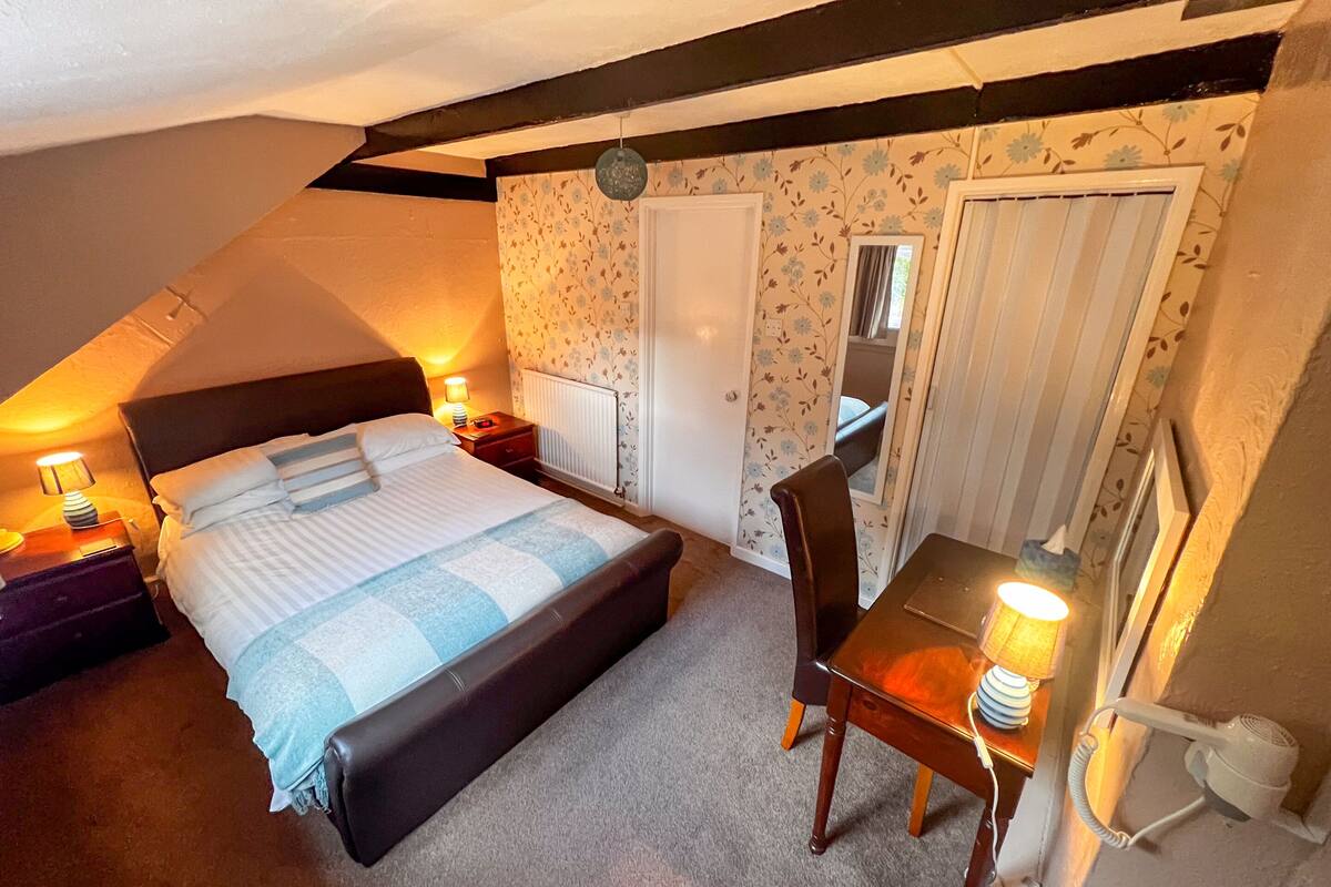 A bedroom with a bed, bedside tables, a desk, and a chair. There is a radiator, a mirror, and a window with curtains.