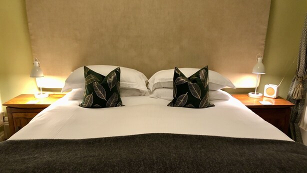 Een keurig opgemaakt bed met witte lakens en twee decoratieve kussens met een bladmotief. Er staan twee nachtkastjes met lampen en een kleine speaker.