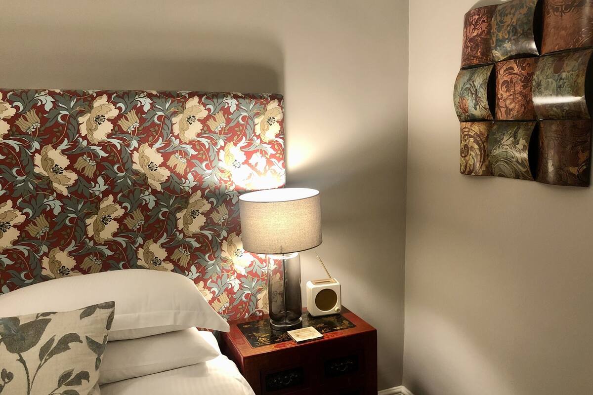 Ein ordentlich bezogenes Bett mit einem Kopfstück mit Blumenmuster, eine Nachttischlampe, ein Radio und ein dekoratives Wandbild.