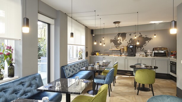 Ein modernes Restaurant mit einer zentralen Sitzgruppe, ausgestattet mit einer blauen Couch und grünen Stühlen, einem Küchenbereich mit einer Weltkarte an der Wand und einem Essbereich mit Marmortischen und -stühlen.