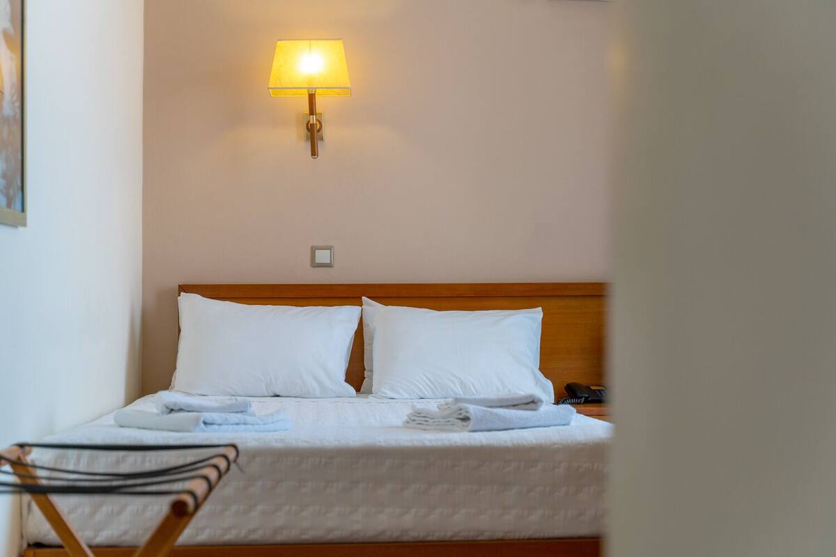 Habitación de hotel con cama, almohadas blancas, lámpara de noche, teléfono y una silla.