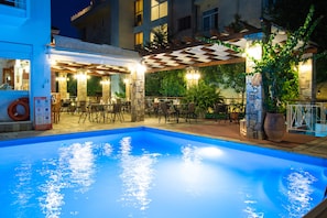 Un área de piscina exterior con una pérgola a cuadros, zonas de estar y un pilar de piedra con una planta.