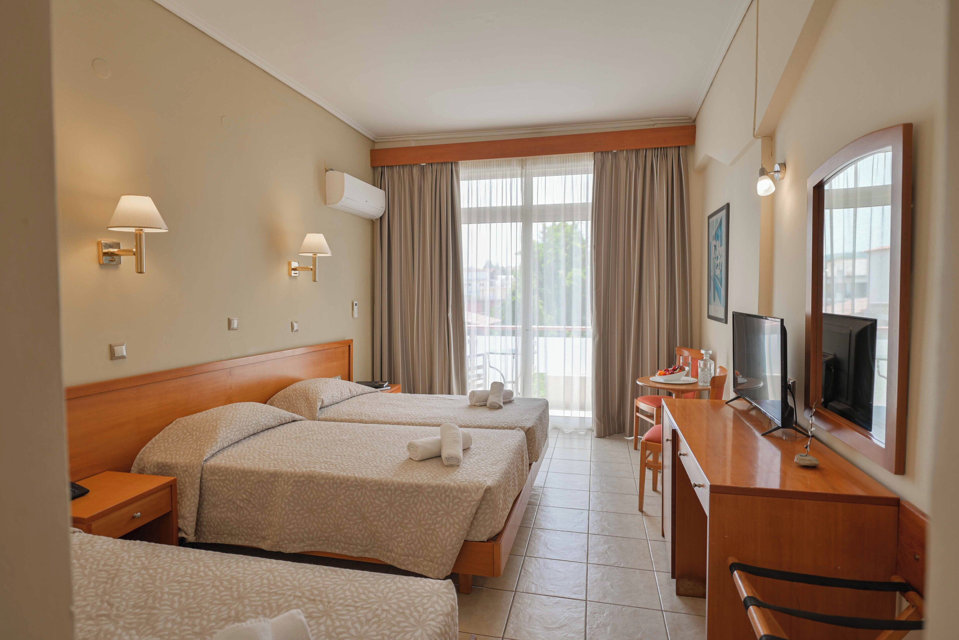 Une chambre d’hôtel avec trois lits, un bureau avec une télévision et un balcon avec des rideaux.