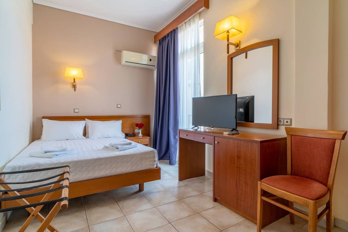 Habitación de hotel con una cama, un escritorio con televisor, una silla y un ventanal con cortinas.