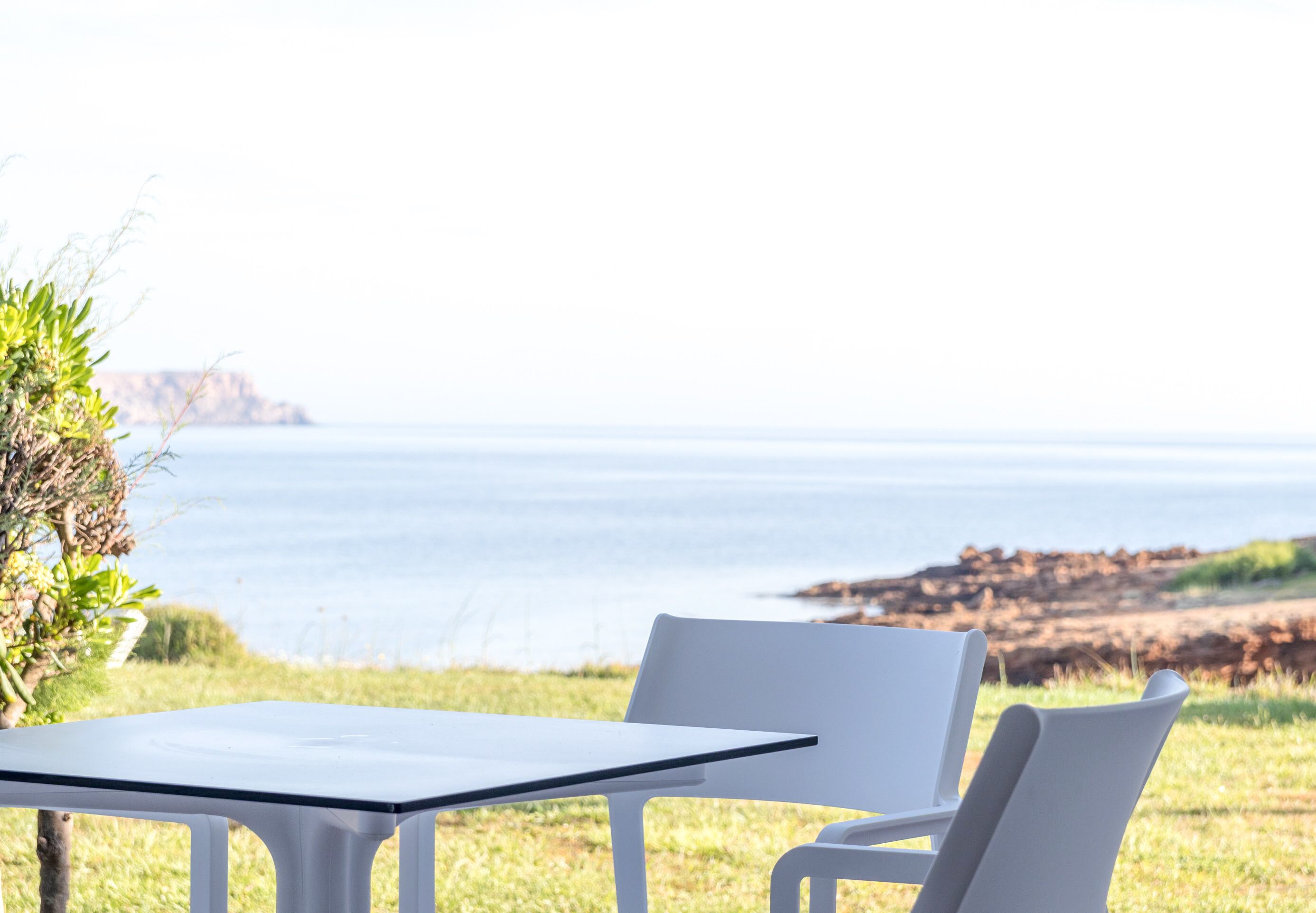 A table and two chairs set outdoors with a view of the sea and rocky coastline.
