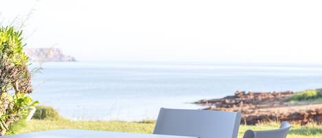 A table and two chairs set outdoors with a view of the sea and rocky coastline.