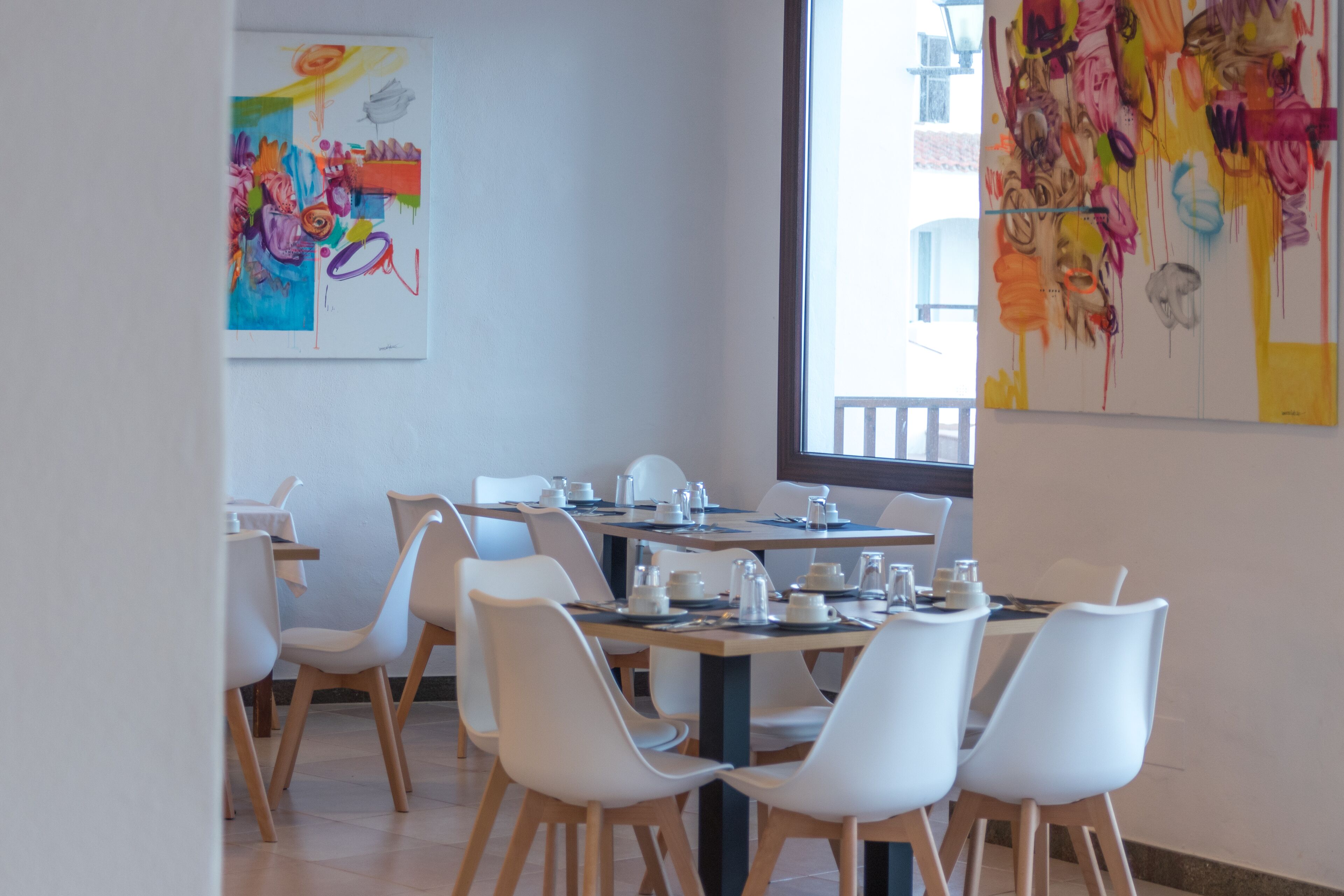 A dining area with white chairs and tables, a large window, and colorful abstract art on the walls.