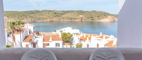 A balcony with white plastic chairs and a table overlooking a coastal town with white buildings and a body of water.