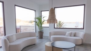 A modern living room with a curved sofa, a round coffee table, and a hanging light fixture. There is a large window providing a view of the sea and a potted plant. - Comitas Tramontana Park (Mercadal)