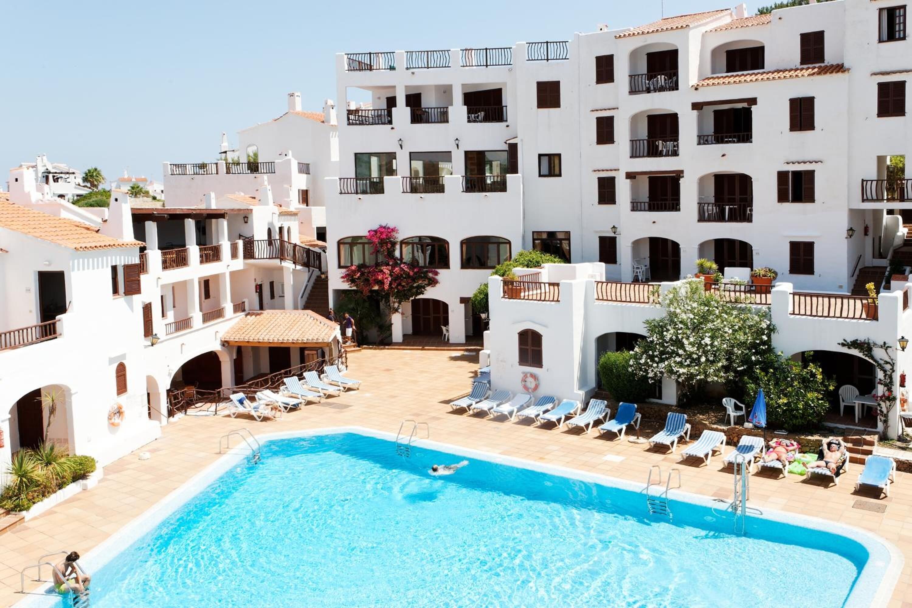 Una piscina rodeada de edificios blancos con balcones, cielo despejado y algunas personas cerca.