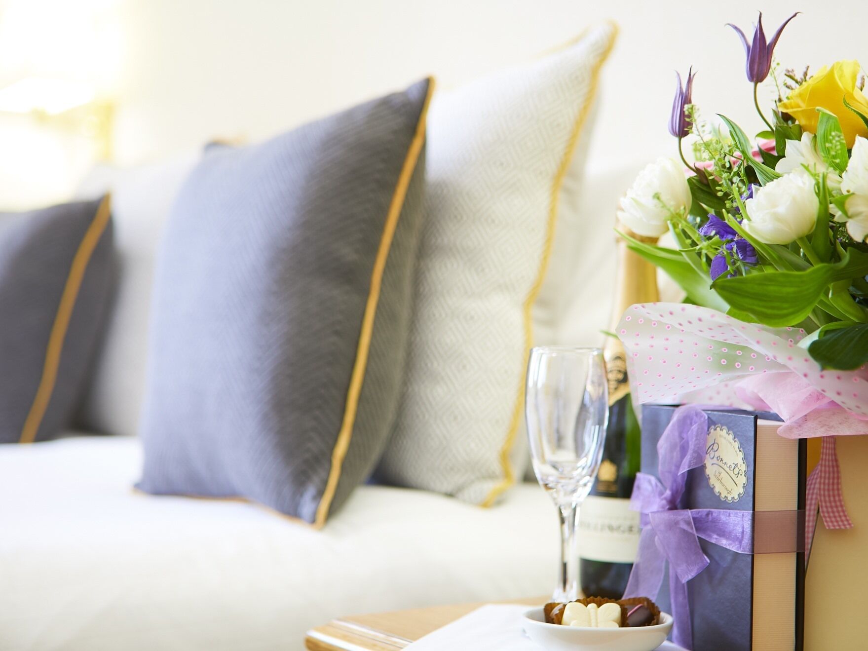 A cozy living room with a white sofa, a glass of wine, a bouquet of flowers, and a gift box.
