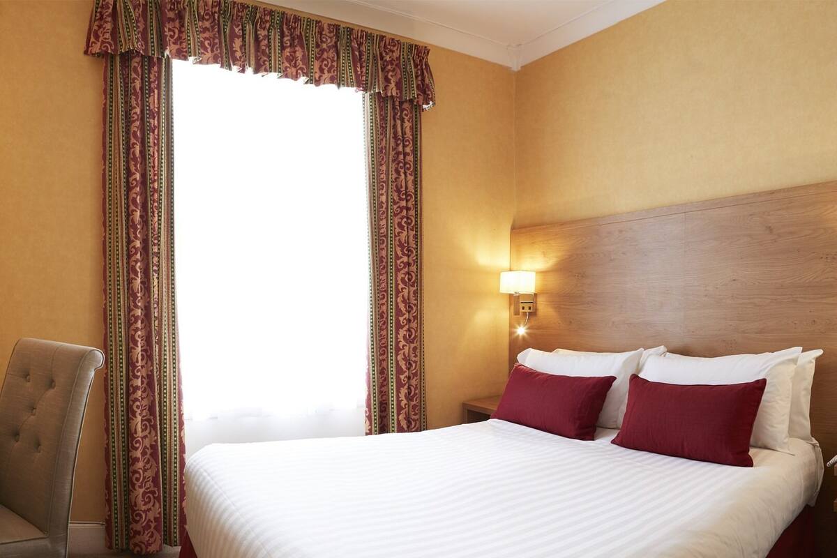 A hotel room with a large bed, two pillows, a nightstand with a lamp, a chair, and a window with curtains.