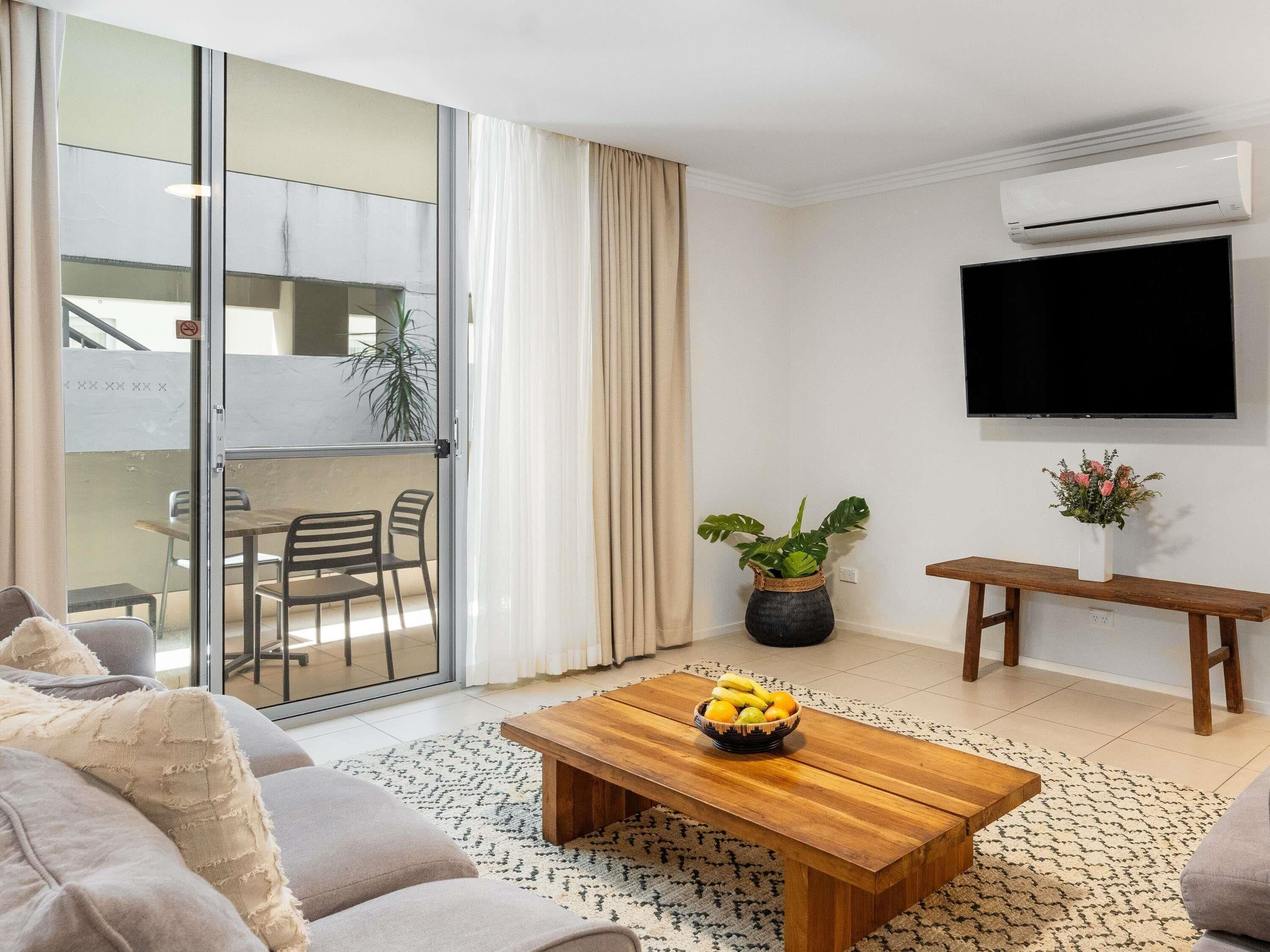 A modern living room with a sofa, a wooden coffee table with fruit, a TV, a plant, and a balcony with a table and chairs.