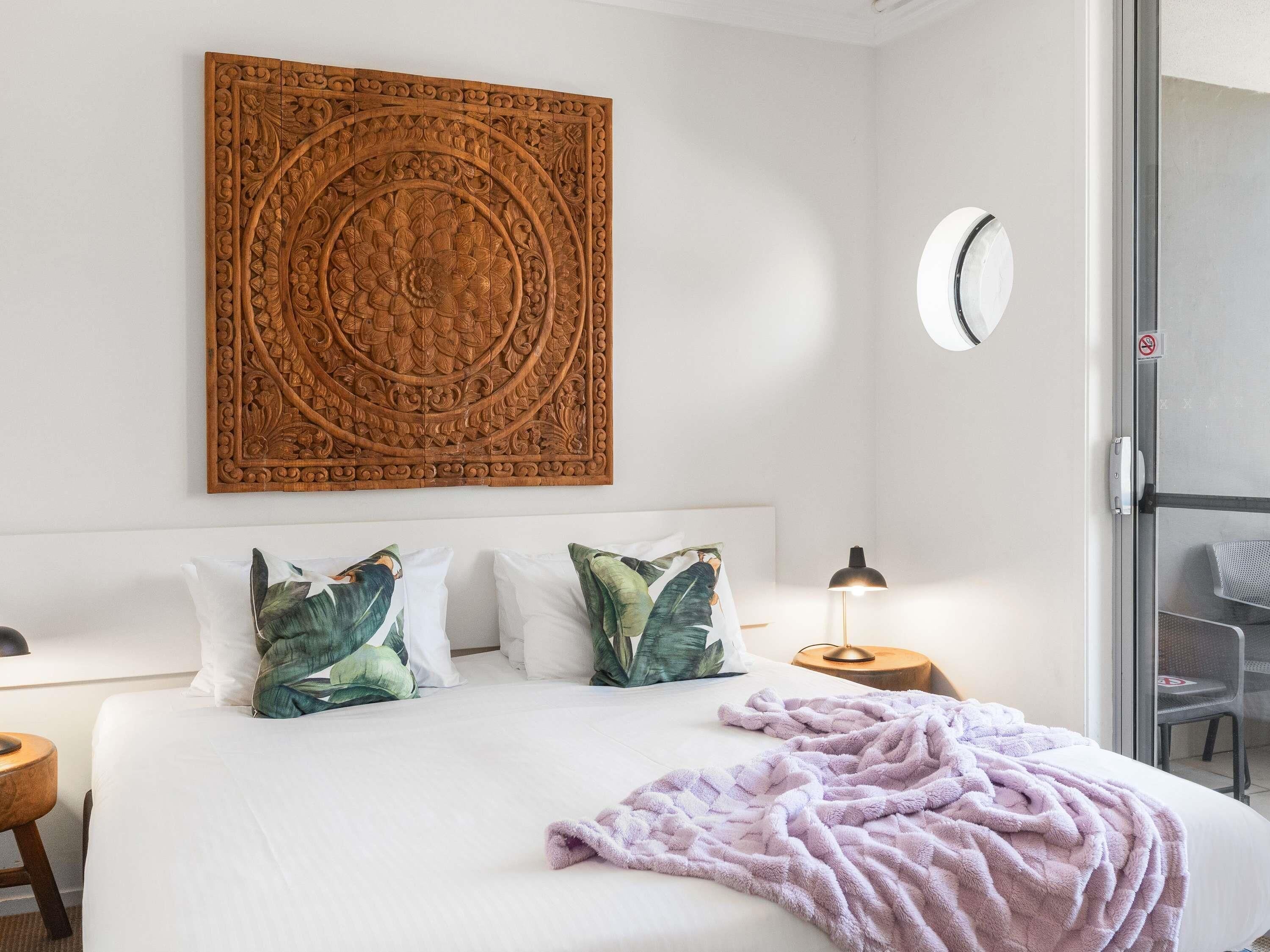 A hotel room with a large bed, two pillows, a wall-mounted decorative panel, a round clock, a nightstand, a lamp, and a sliding glass door leading to a balcony.