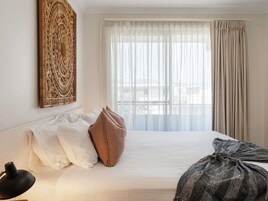 Una habitación de hotel con una cama grande, almohadas blancas, una almohada decorativa marrón, una lámpara negra y un ventanal con cortinas translúcidas.