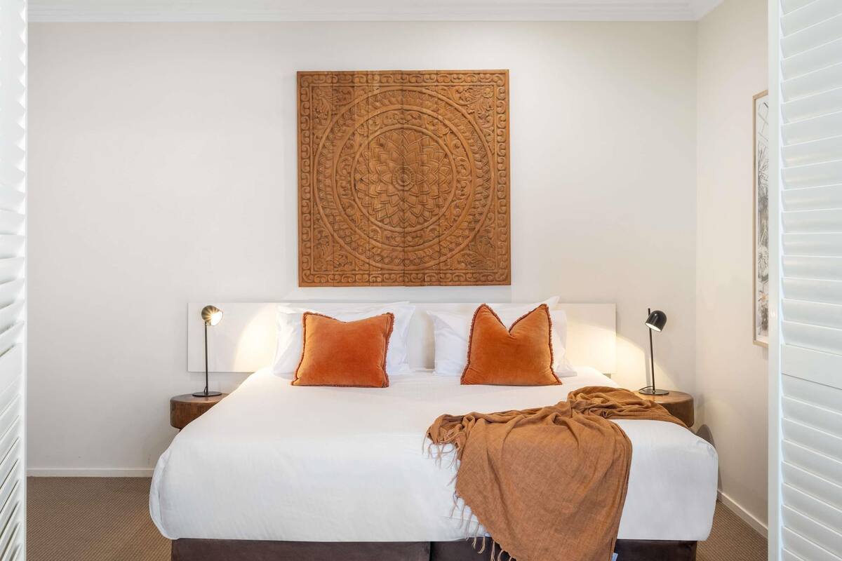 A neatly made bed with white linens and two orange pillows, a decorative wall piece above, and a bedside lamp on each side.