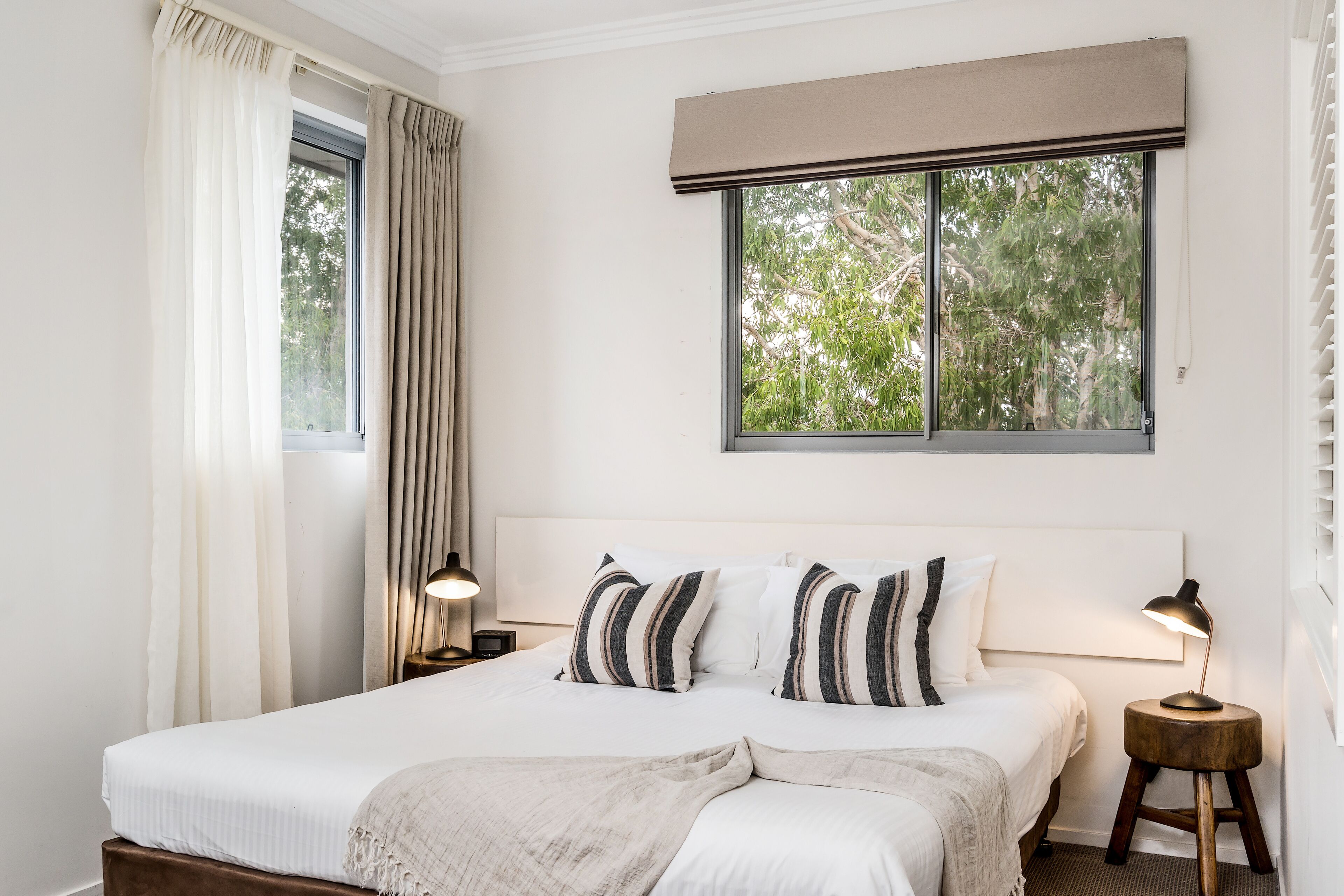 A bedroom with a large bed, two bedside lamps, a window with a view of trees, and a headboard with a light-colored shade.