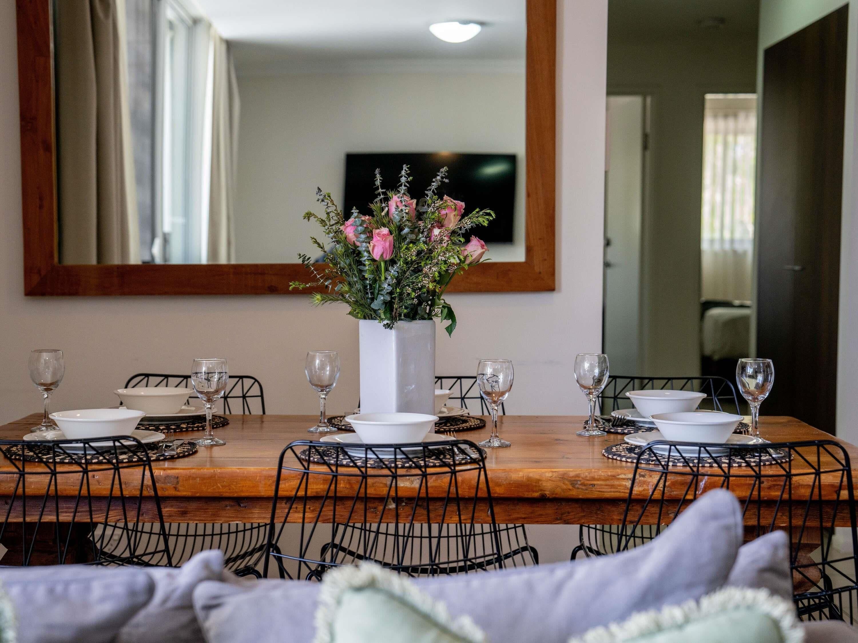 A dining table set with white plates, bowls, and wine glasses, surrounded by black wire chairs, with a large mirror above and a floral centerpiece.