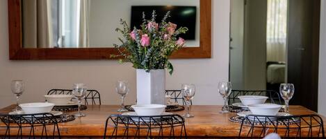 Une table à manger dressée avec de l’assiette, des bols et des verres à vin blancs, entourée de chaises en fil de fer noir, avec un grand miroir au-dessus et un centre de table floral.