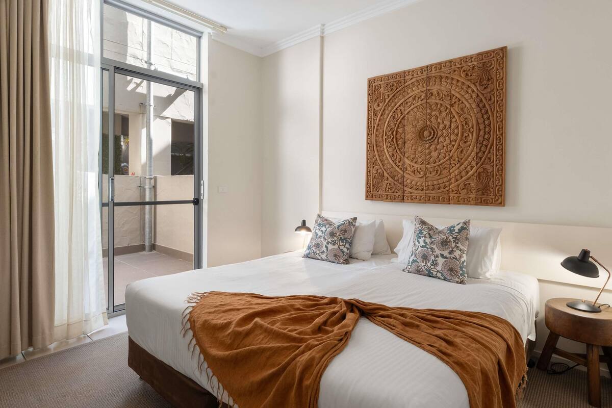 A hotel room with a large bed, a patterned wall hanging, a small bedside table with a lamp, and a sliding glass door leading to an outdoor area.