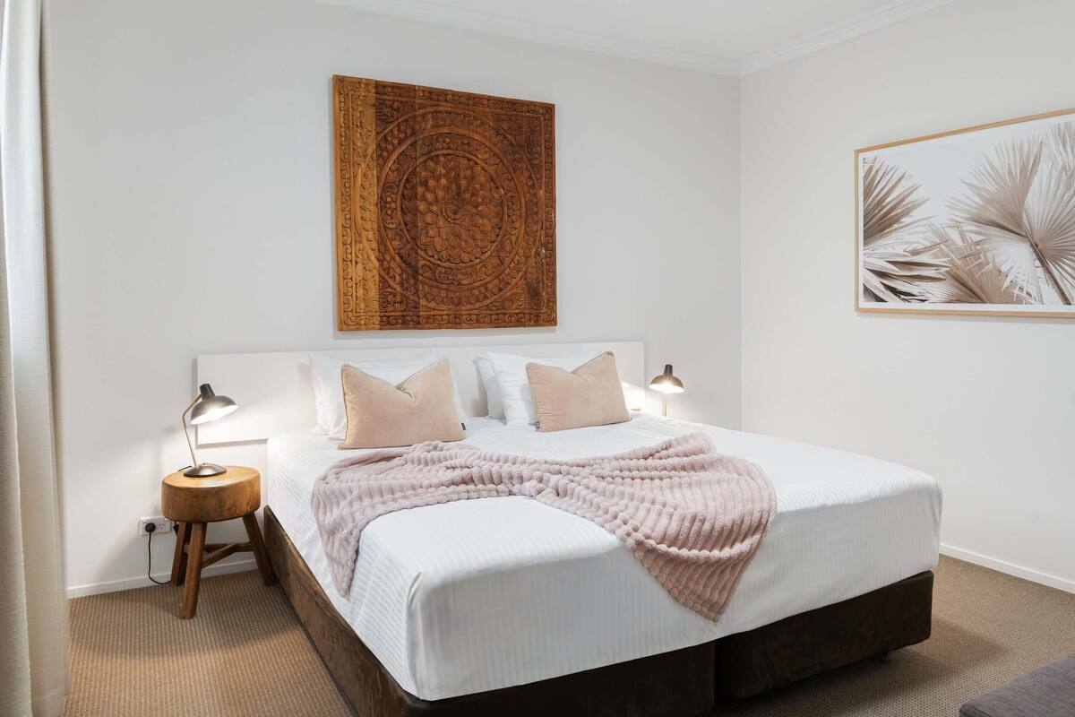 A neatly arranged bedroom with a large bed, bedside lamps, a decorative wall piece, and a framed artwork.