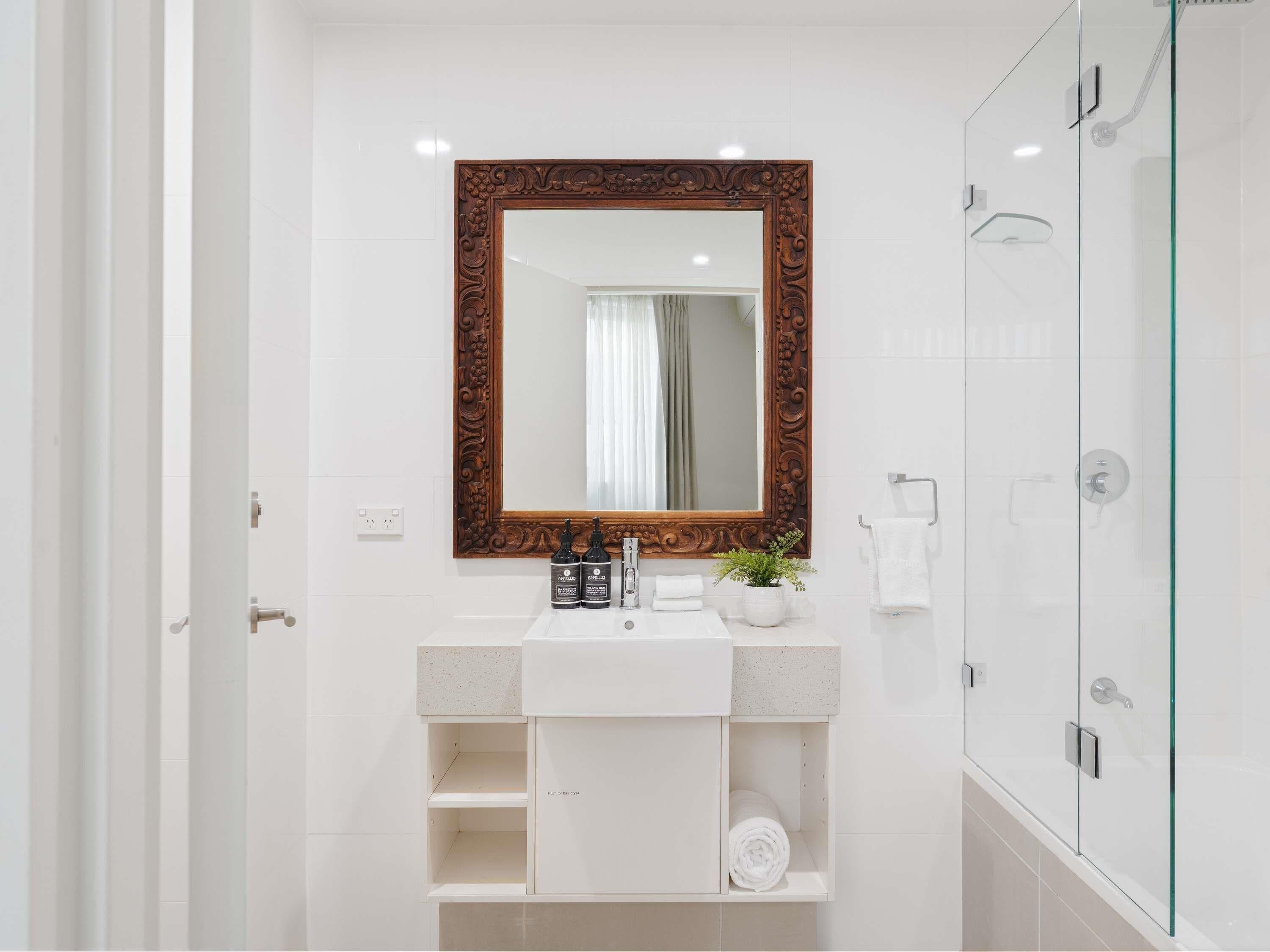 A modern bathroom with a large mirror, a white sink, a glass shower enclosure, and a potted plant.