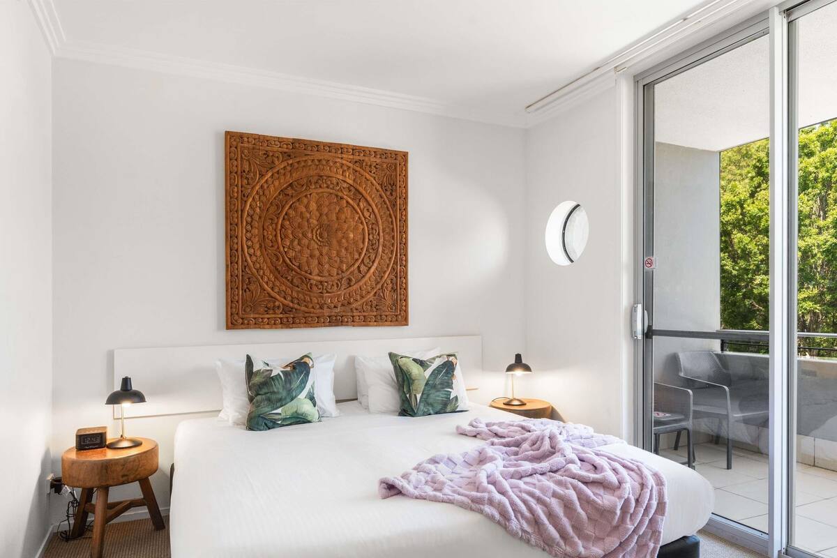 A bedroom with a large bed, a decorative wall piece, a bedside table with a lamp, and a view of the outdoors through a sliding door.