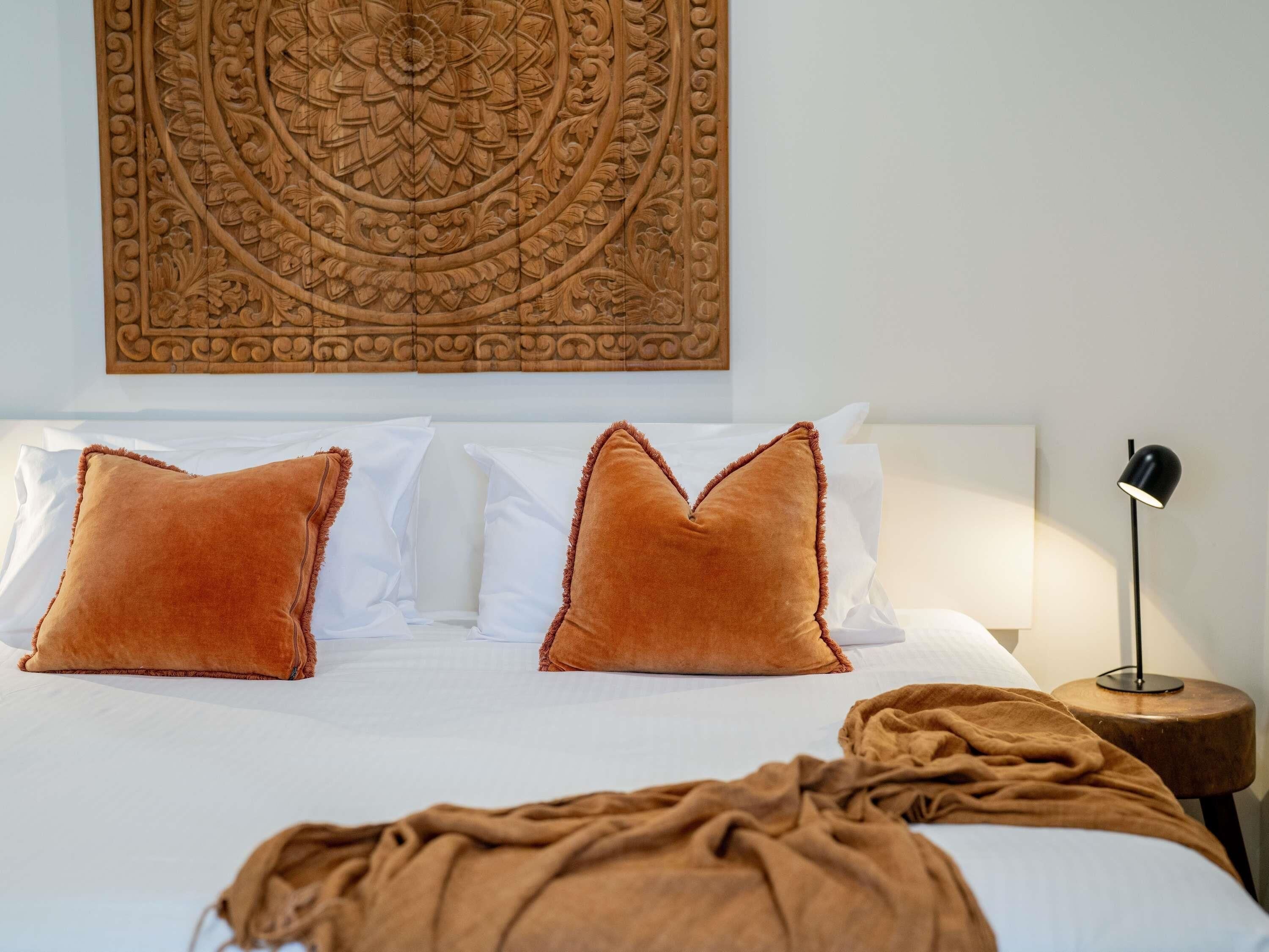 Una cama con sábanas blancas y dos almohadas marrones, una manta color café y un tapiz de pared, junto a una lámpara de noche.