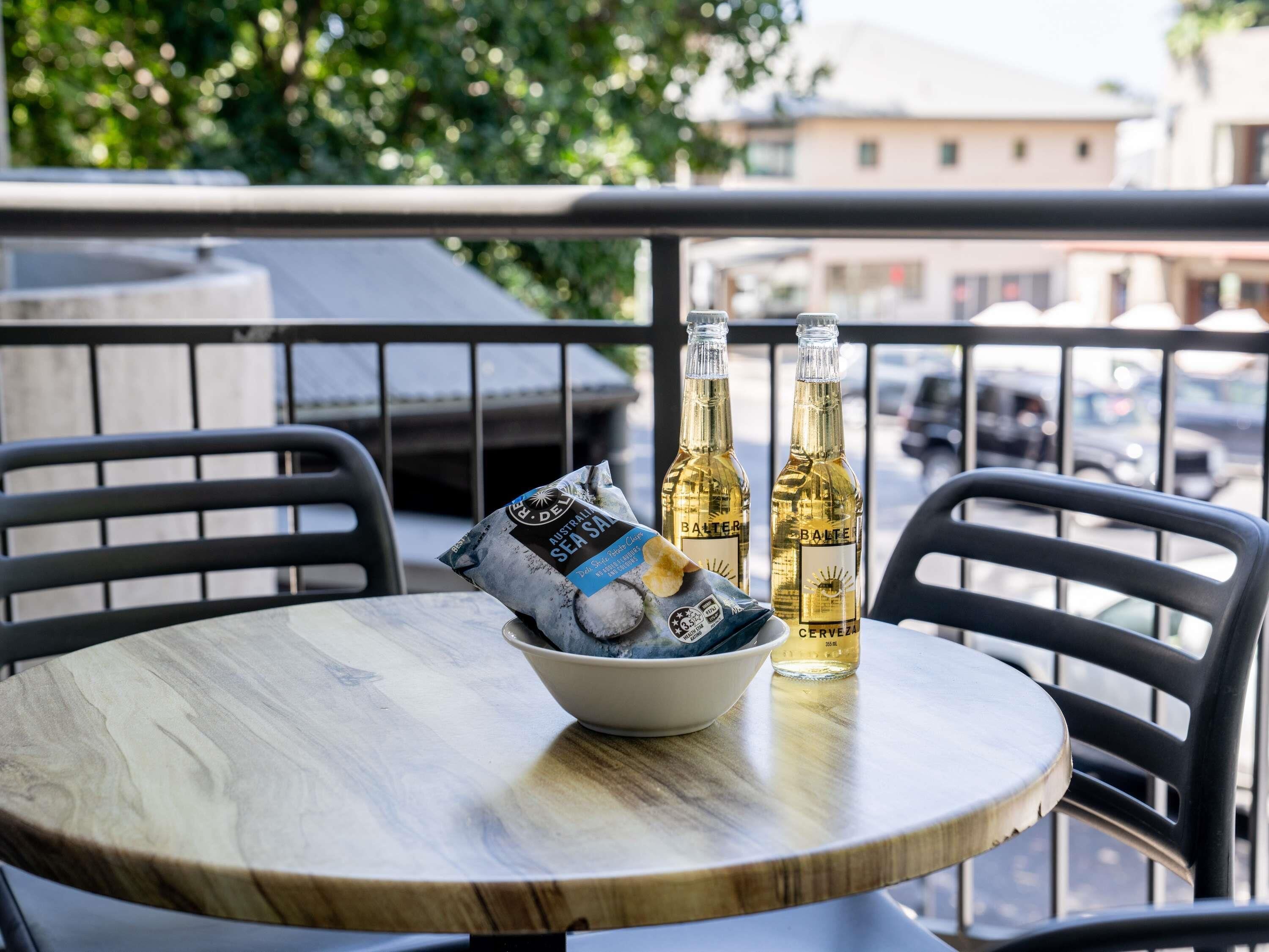 A round wooden table with a bowl of snacks and two bottles of beer on a balcony.
