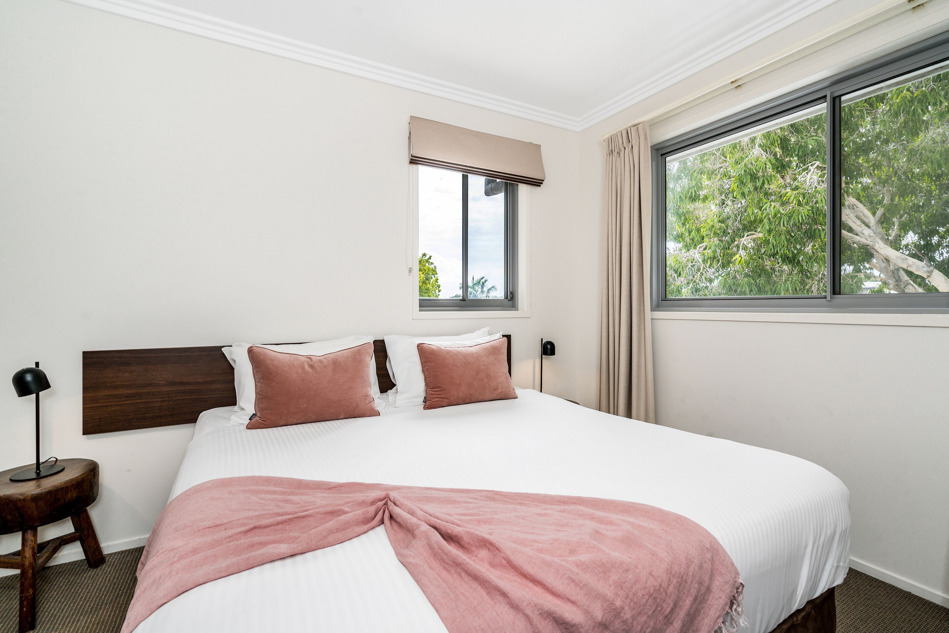 A neatly arranged hotel room with a large bed, a bedside table with a lamp, a window with curtains, and a view of trees outside.