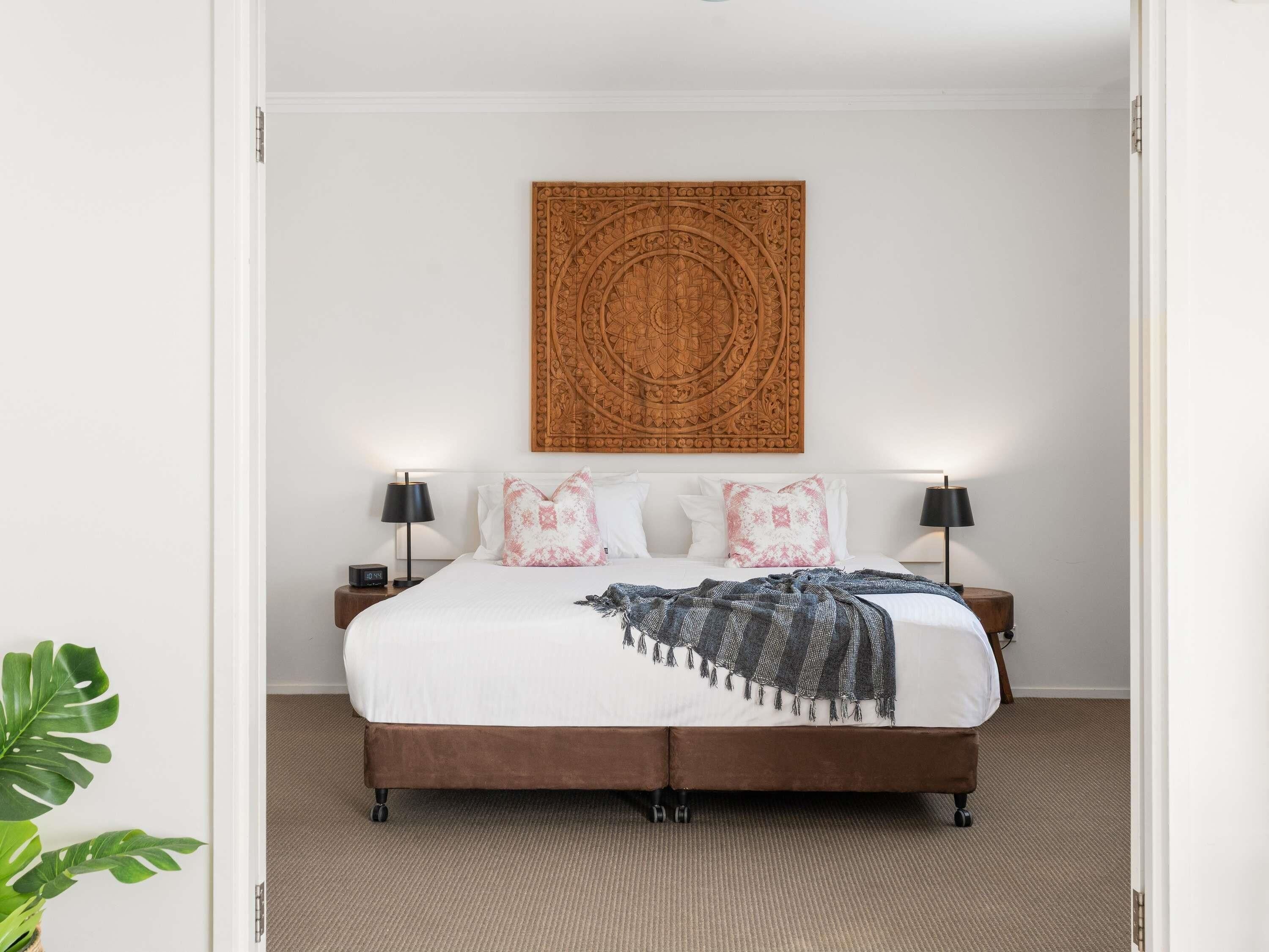 A bed with a brown headboard, white bedding, and decorative pillows. There is a wall-mounted artwork above the bed.