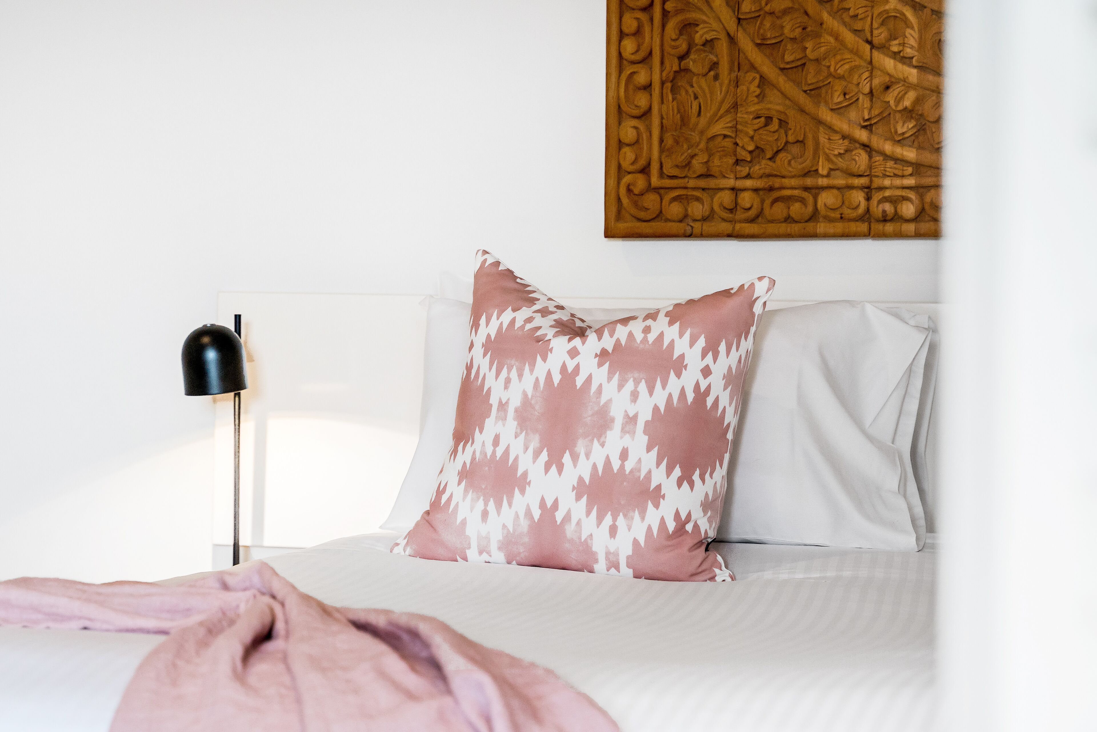 Una cama bien hecha con almohadones estampados, una lámpara de noche y un adorno de pared decorativo.