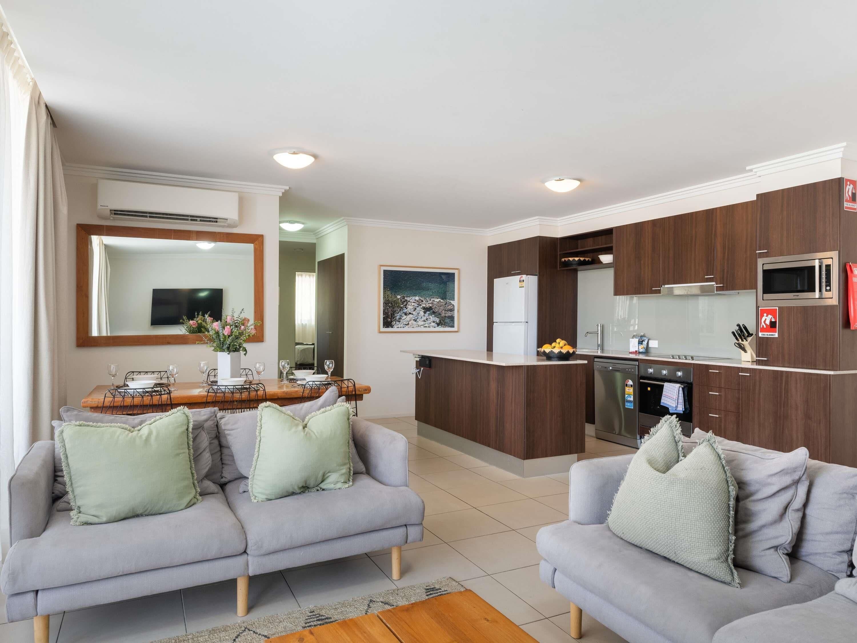 A modern living room with two grey sofas, a wooden dining table, and a kitchen with dark wood cabinets and stainless steel appliances.