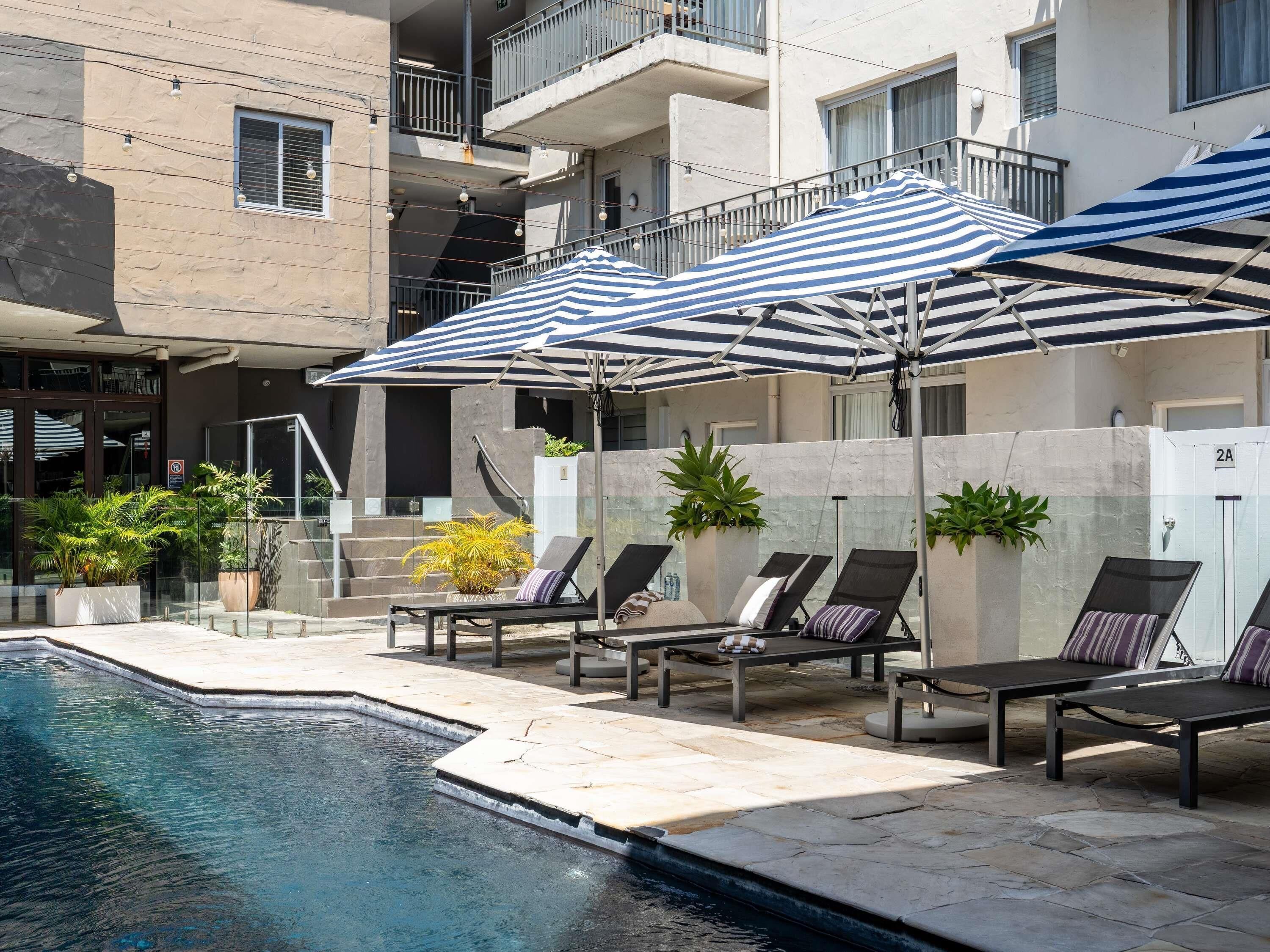 A poolside area with lounge chairs and striped umbrellas, a stone pool, and a building with balconies.