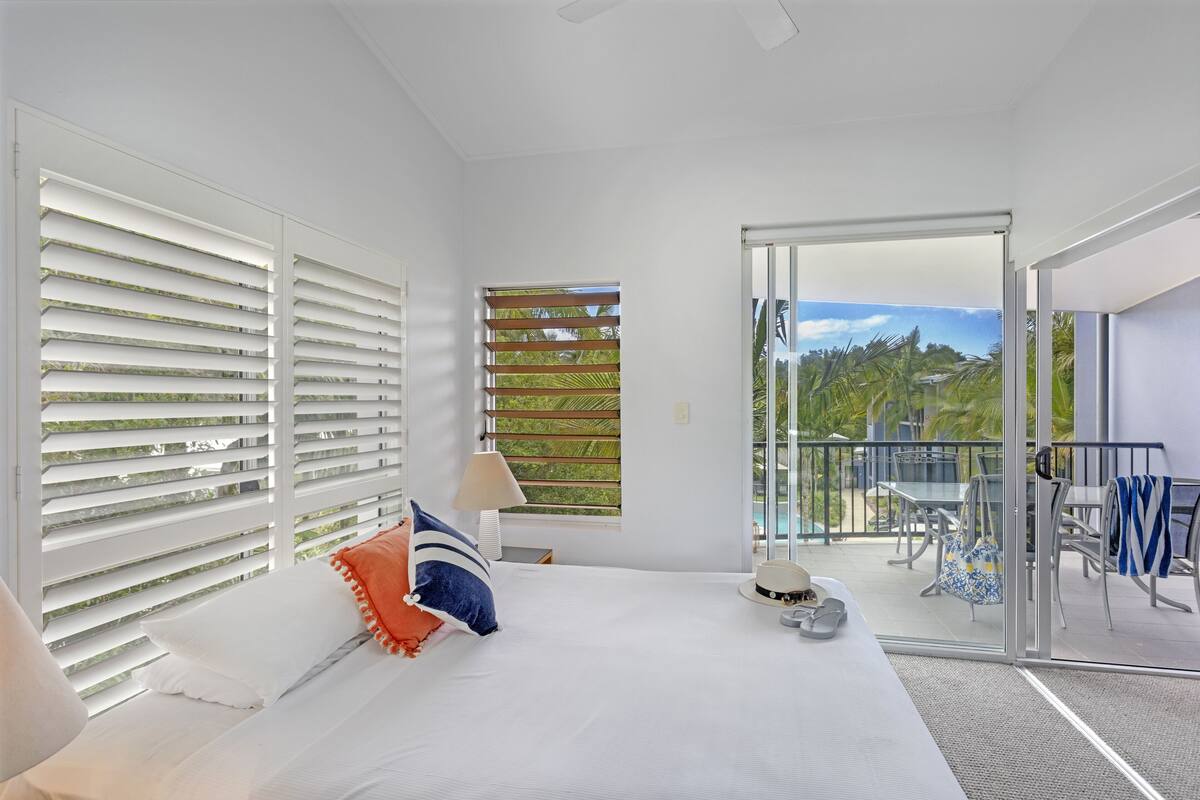 A bedroom with a bed, a window with blinds, a balcony with outdoor furniture, and a view of greenery.