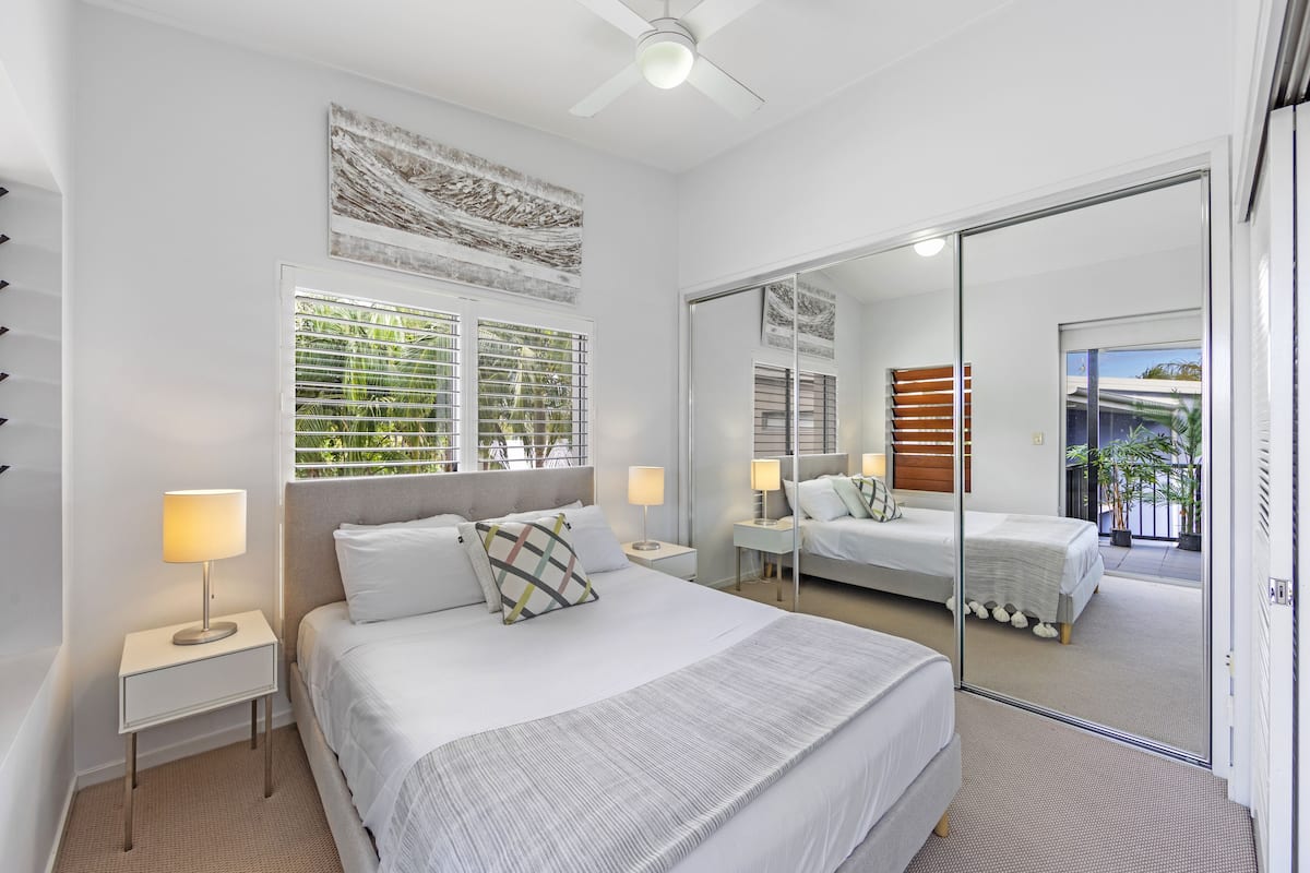 A modern bedroom with a large bed, bedside tables with lamps, a ceiling fan, and a sliding glass door leading to a balcony.