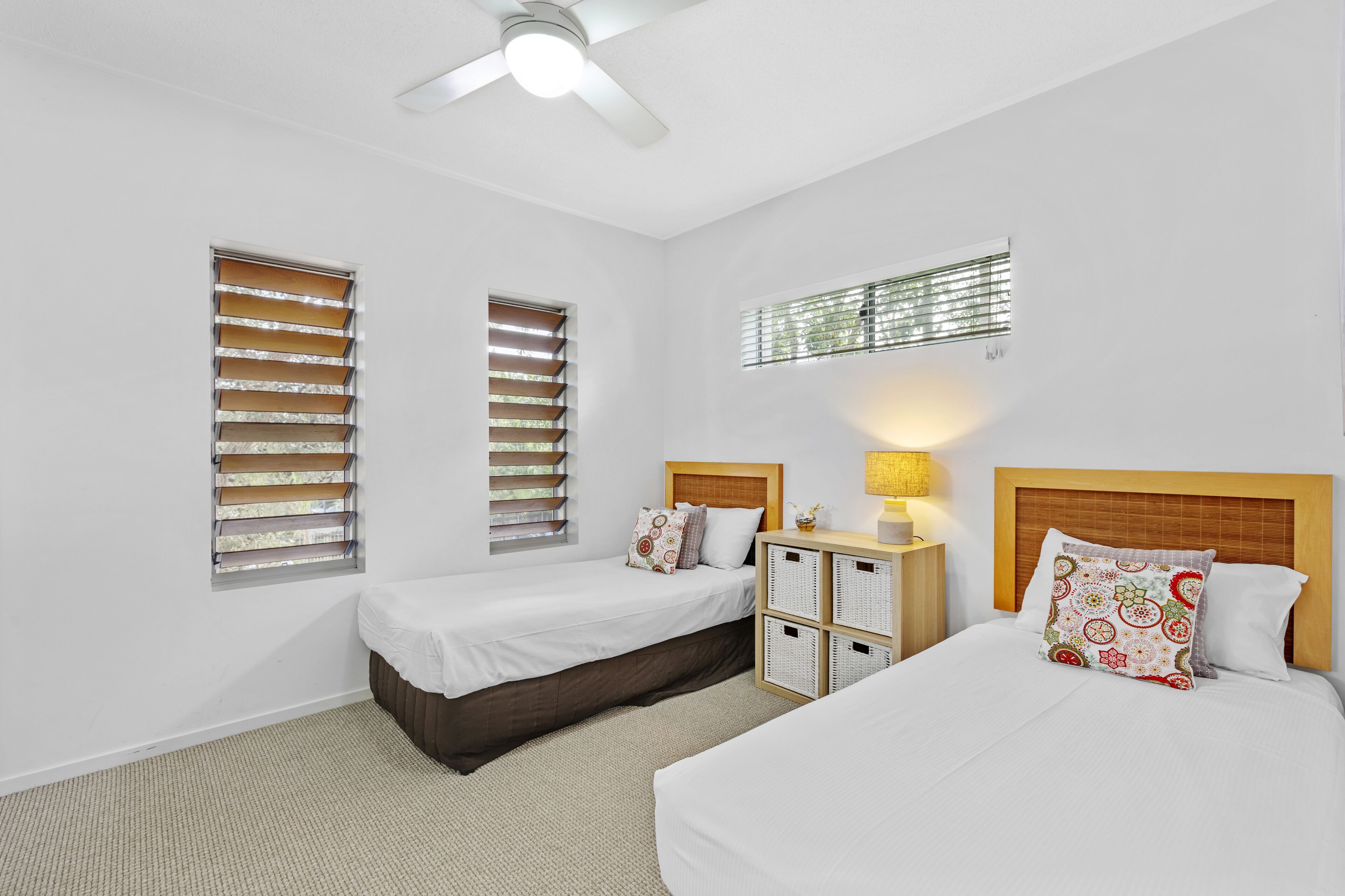 A hotel room with two beds, a ceiling fan, a window with blinds, a nightstand with a lamp, and a headboard with a decorative pillow.