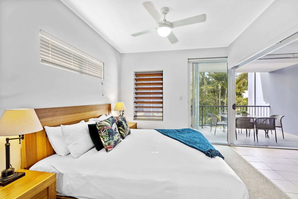 A modern hotel room with a large bed, a ceiling fan, a balcony with outdoor furniture, and a view of palm trees.
