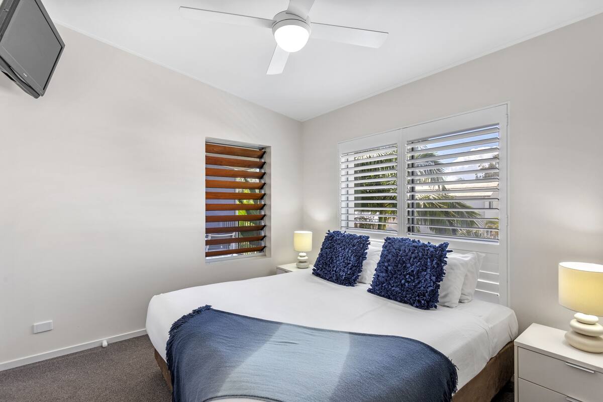 A bedroom with a bed, two bedside lamps, a ceiling fan, and a window with blinds.