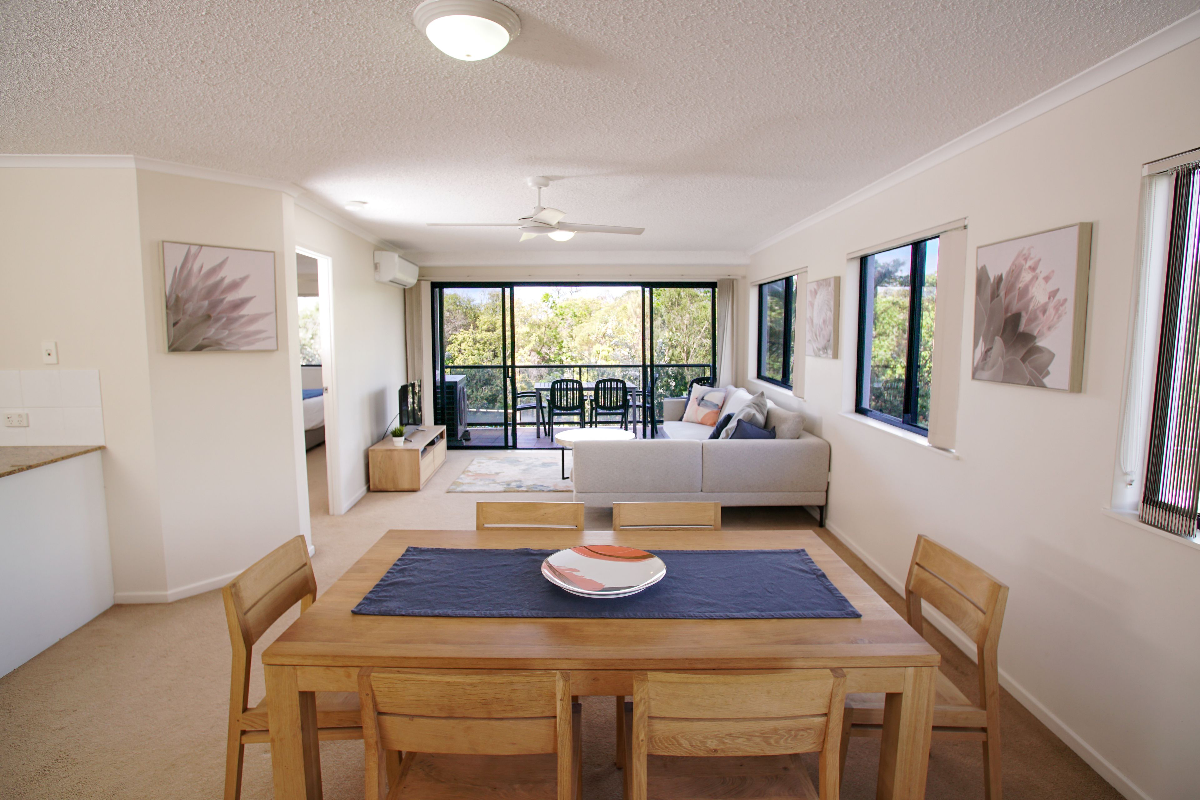 A dining area with a wooden table, chairs, and a blue placemat. There is a sofa and a window with a view of trees in the background.