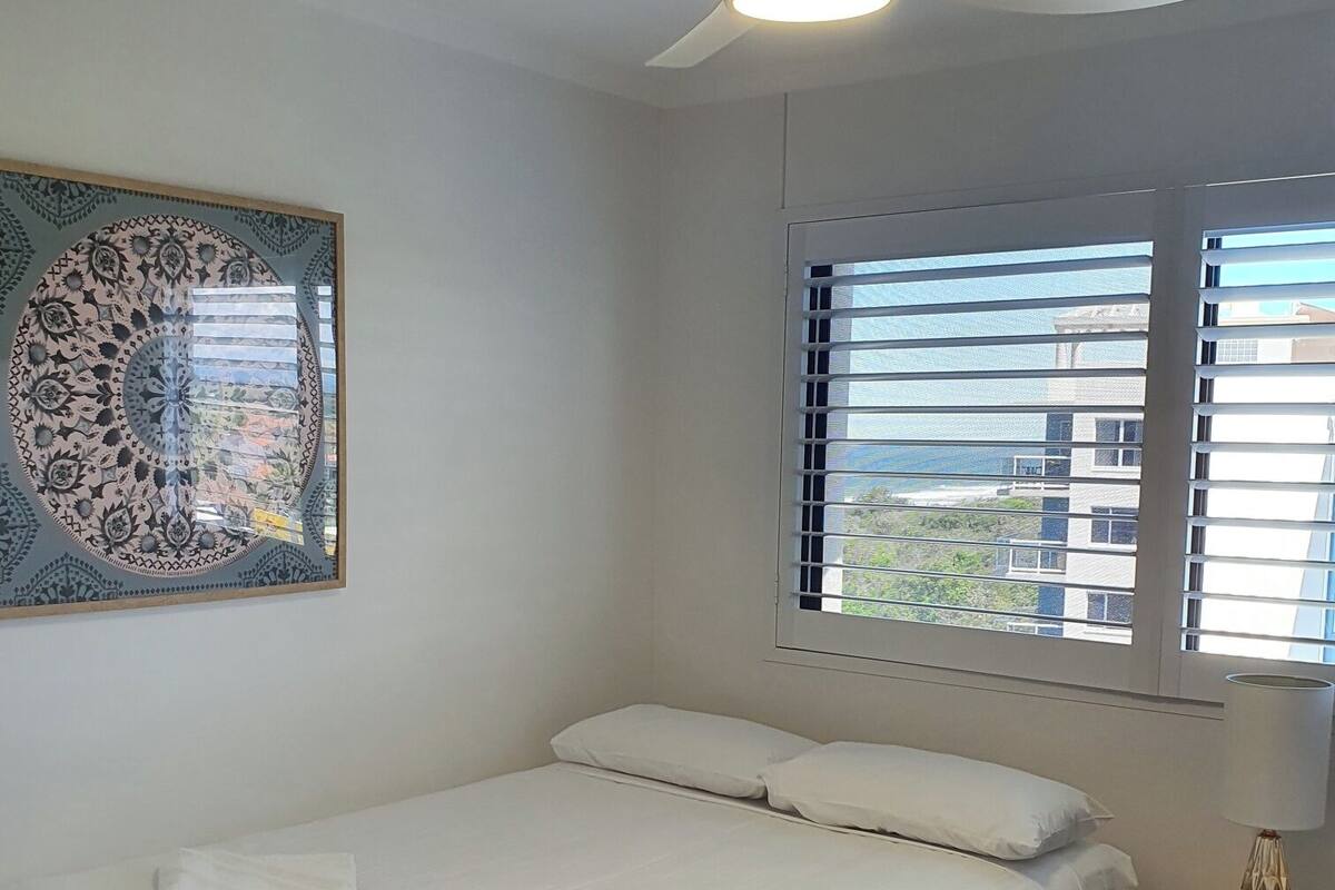 A bedroom with a bed, a ceiling fan, a window with blinds, and a wall-mounted artwork.