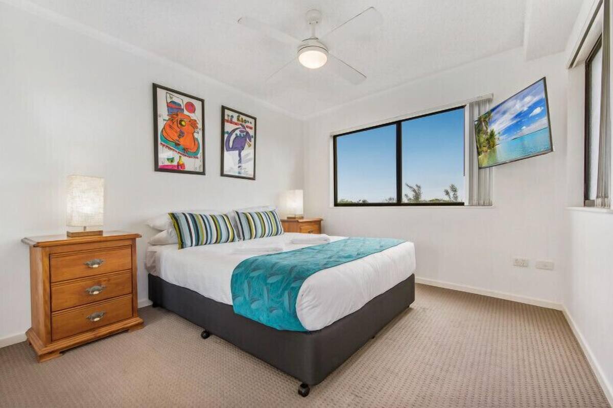 A bedroom with a large bed, a wooden nightstand, a ceiling fan, and a window with a view of the beach.